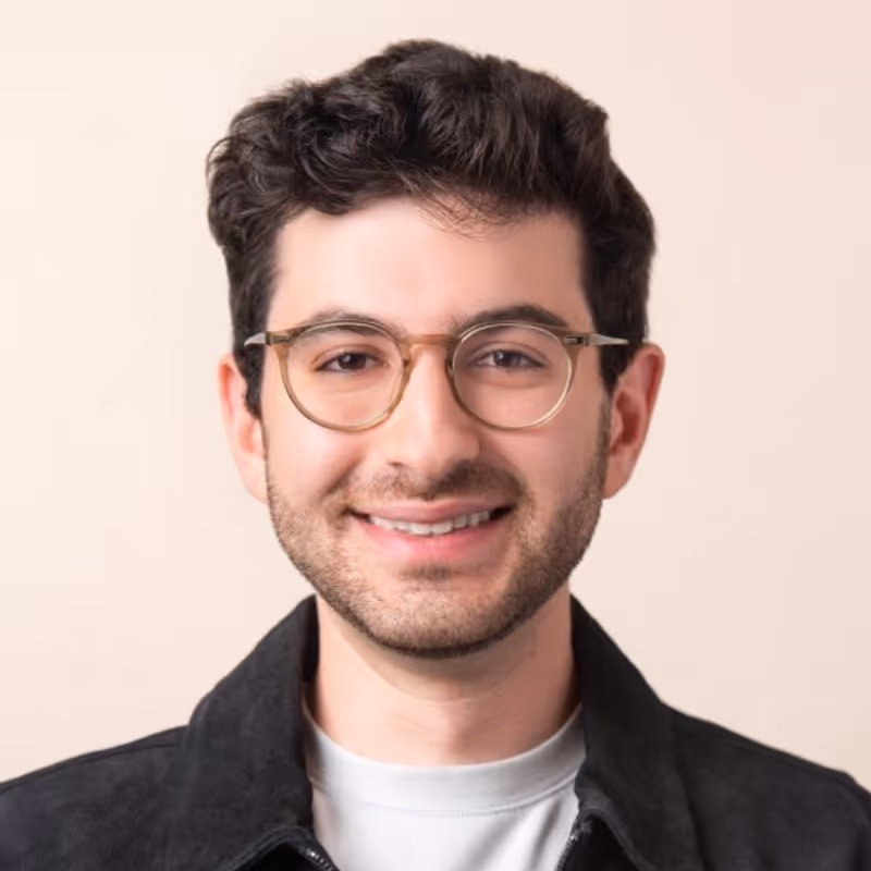 Young man with curly dark hair, wearing glasses and a black jacket, smiling against a beige background.